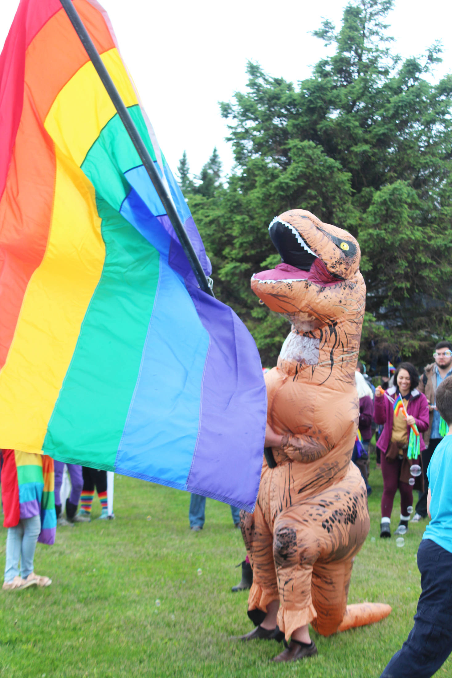 Gage Dillon, 14, waves a pride flag while dressed in a dinosaur suit before Homer’s first ever Pride March on Saturday, June 23, 2018 at WKFL Park in Homer, Alaska. About 280 people filled the streets during the march from the park to Grace Ridge Brewery. (Photo by Megan Pacer/Homer News)