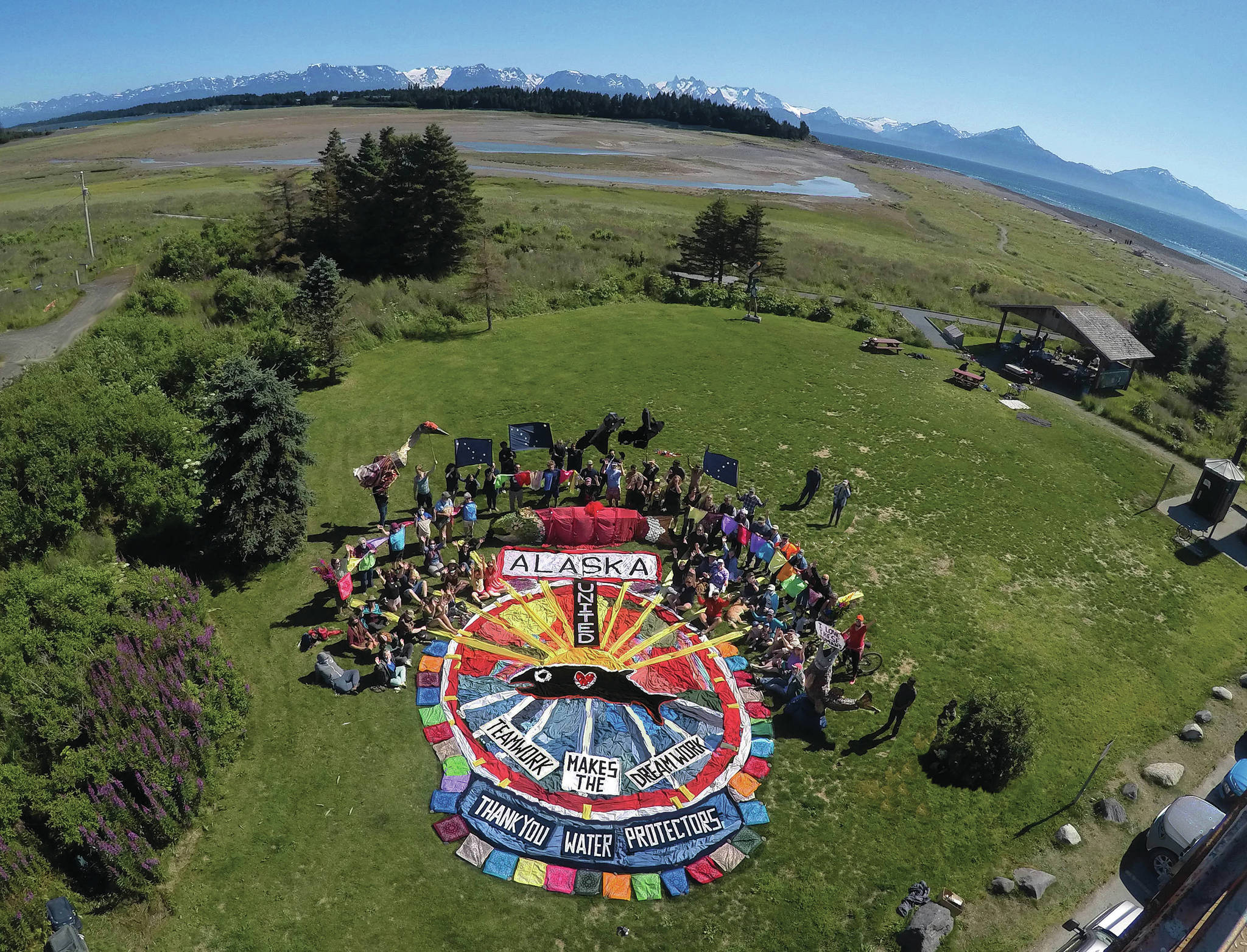 Homer’s Bishop’s Beach Park was the location for an enactment of public participatory art on Sunday, Aug. 1, 2021, in Homer, Alaska. A ground design of colored fabric was created to convey a message of “Alaska United,” “Teamwork Makes the Dream Work” and “Thank You Water Protectors.” The public was invited to be part of the art by standing and sitting around the central design. Salmon sculptures, raven and sandhill crane puppets, Alaska flags and colored bandanas were an added touch to the overhead image depicting salmon solidarity.  The project was led by Mavis Muller and was the finale to her series of 12 annual aerial group photos for the protection of Alaska’s Bristol Bay. Photographer Russell Campbell captured the photo from a bucket lift at 35 feet high.
“Art is communication. With our creativity and imagination we can inspire new possibilities, and we can have fun doing it,” Muller said. (Photograph by Russell Campbell)